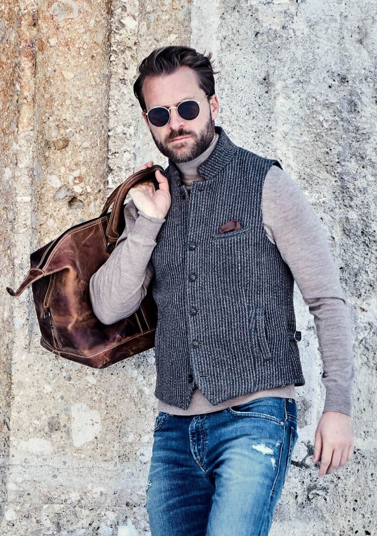 Model trägt DORNSCHILD Herrenweste HW26 W11-1 in Grau-Blau mit Stehkragen, Rollkragenpullover, Jeans und Lederreisetasche
