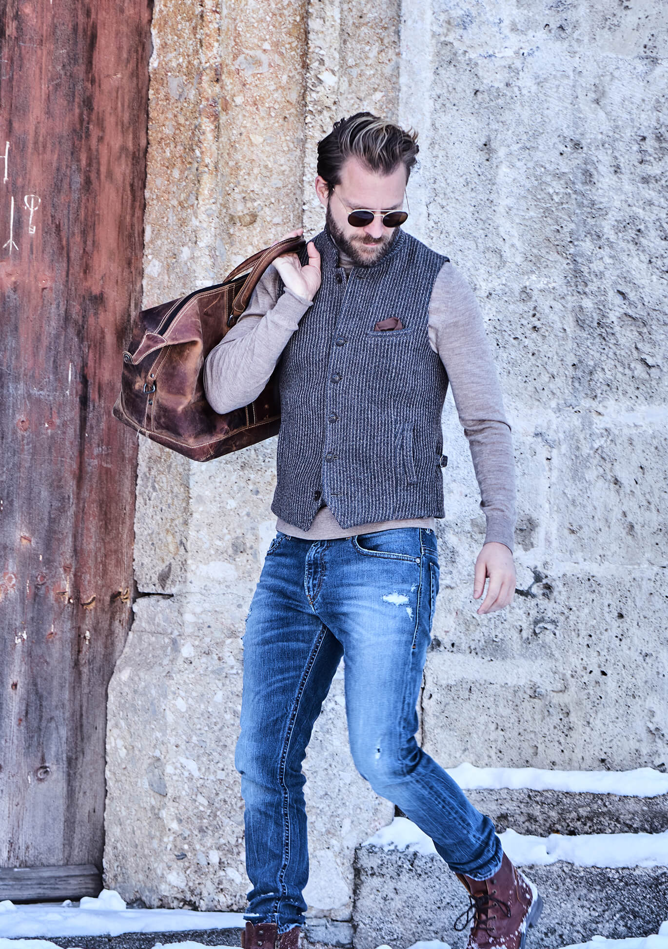 Model trägt DORNSCHILD Herrenweste HW26 W11-1 in Grau-Blau mit Stehkragen, Rollkragenpullover, Jeans und Lederreisetasche